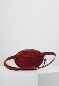 Borsa ovale in pelle rossa con una texture liscia, sottile tracolla regolabile e chiusura con zip, con due accenti orizzontali sul retro.