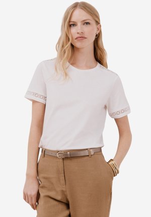 T-shirt blanc à manches courtes avec dentelle sur les manches, col rond, associé à un pantalon beige taille haute et une ceinture assortie.