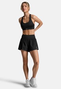 Zwarte sportbeha en zwarte high-waisted shorts met een uitlopende zoom. Model staat in witte sneakers tegen een effen achtergrond.