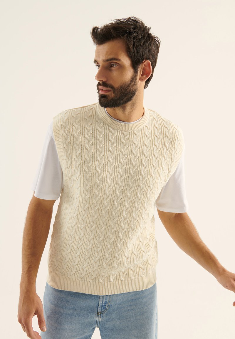 Vesta din tricot bej deschis, cu model cu ochiuri, cu decolteu rotund, purtată peste o cămașă albă cu mânecă scurtă, asortată cu blugi albaștri deschiși.