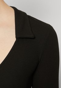Svart ribbet stofftopp med en bred, flat krage og en dyp V-hals, som viser en jevn tekstur og en tettsittende design.