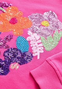 Tissu rose vif avec des motifs floraux colorés en sequins et perles, et une manche à revers côtelée visible dans le coin.