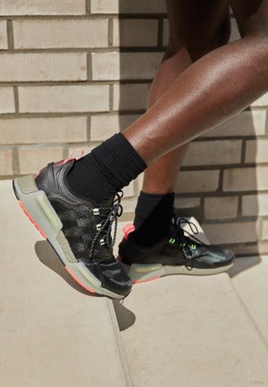 Gambe con calzini neri e scarpe da ginnastica nere con motivo che scendono scale di cemento chiaro accanto a un muro di mattoni.