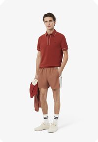 Camisa polo roja con detalles blancos, pantalones cortos beige con paneles laterales, zapatillas beige y calcetines blancos con rayas negras.