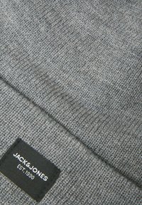 Beanie grigio in maglia con un motivo testurizzato. Presenta un'etichetta nera con testo bianco nell'angolo in basso. Materiale morbido e vestibilità aderente.