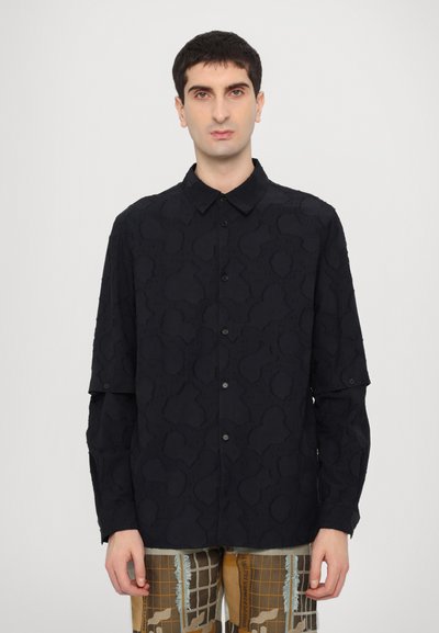 Schwarze Hemdjacke mit strukturiertem Design, mit Kragen und langen Ärmeln. Getragen mit gemusterten Hosen.