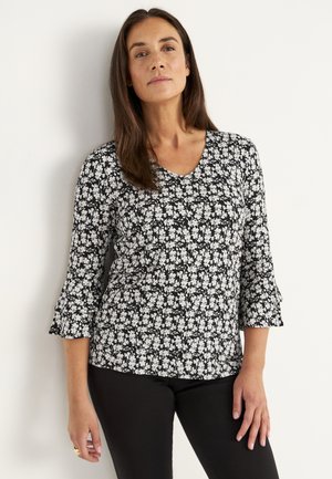 Vrouw met lang bruin haar in een zwart-witte bloemmotief V-hals top met 3/4 uitlopende mouwen en zwarte broek, staand tegen een witte muur.