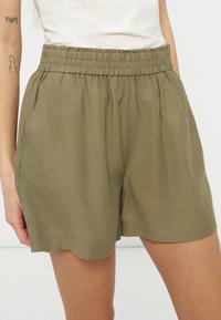 Γυναίκα που φοράει λινά σορτς με ελαστική μέση σε ελιά χρώμα, με πλαϊνές τσέπες και λευκό τοπ, εμφανίζοντας μήκος μέχρι τα μισά του μηρού και άνετη εφαρμογή.