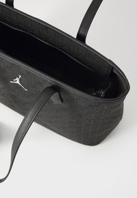 Schwarze Tragetasche mit geprägtem Muster, ausgestattet mit zwei Griffen und einem silbernen Logo. Das Innere zeigt ein strukturiertes Futter und ein offenes Design.