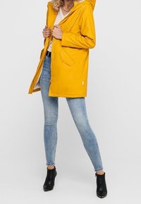 Veste imperméable jaune avec capuche, fermeture à boutons-pressions et zip, équipée de poches latérales. Portée sur un haut léger et un jean skinny. Bottines noires.