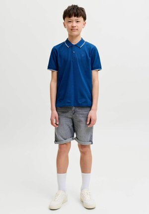 Gutt står med nøytral ansiktsuttrykk iført blå polotrøye, grå oppbrettede shorts, hvite sokker og hvite joggesko på hvit bakgrunn.