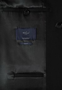 Intérieur de veste noire slim fit avec étiquette bleu marine indiquant la taille et fabriquée au Bangladesh, bouton visible et poche intérieure.