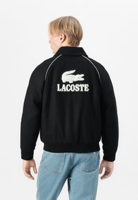 Jachetă neagră cu logo-ul Lacoste brodat alb și crocodil pe spate. Are guler, mansete elasticizate și o textură netedă.