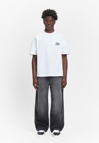 Weiße Baumwoll-T-Shirt mit kurzen Ärmeln, ausgestattet mit einem blauen Grafiklogo auf der linken Brust. Kombiniert mit locker sitzenden grauen Jeans aus Denim und weißen Sneakers.