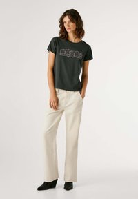 Camiseta negra de manga corta con un gráfico de "Pepe Jeans" en letras texturizadas, combinada con pantalones rectos beige y zapatos de cuña negros.