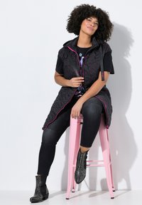 Chaleco negro acolchado con acentos rosas, usado sobre una camiseta gráfica negra, combinado con jeans oscuros y botas de tobillo con tachuelas, sentado en un taburete rosa.