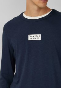 Maglietta a maniche lunghe di colore blu navy con scollatura rotonda. Presenta una toppa bianca con la scritta "Iscriviti alla Natura". Tessuto morbido e leggero.