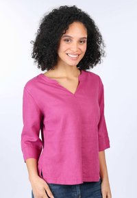 Rosa Leinenbluse mit V-Ausschnitt, Dreiviertelärmeln und seitlichen Schlitzen, getragen mit blauen Jeans. Glatte Textur mit einem subtilen Glanz.