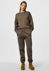 Felpa marrone e pantaloni jogger coordinati realizzati in morbido tessuto. Presenta un colletto a girocollo, spalle abbassate e polsini ed elastico in vita.