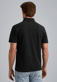 Zwarte polo, korte mouwen, zachte stof, kraag en een klein logo op de rug. Draag het met lichtblauwe jeans voor een casual look.