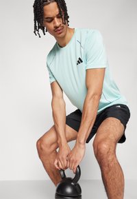 Lys blå treningsskjorte med korte ermer og svart logo. Båret med svarte shorts, holder personen en kettlebell, og viser frem muskuløse armer.