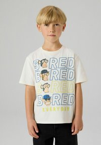 Blond jongetje met een wit T-shirt met cartoonapen gezichten en grote tekst "BORED EVERY DAY" in blauw en geel, zwarte broek.