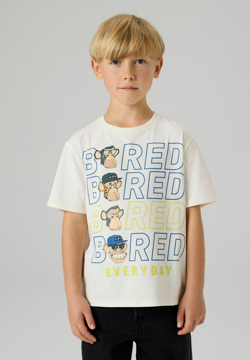 Blond jongetje met een wit T-shirt met cartoonapen gezichten en grote tekst "BORED EVERY DAY" in blauw en geel, zwarte broek.