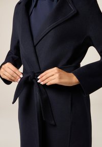 Cappotto di lana blu navy, con cintura, colletto a scollatura e maniche lunghe, dal texture morbida. I dettagli delle cuciture visibili arricchiscono il design.