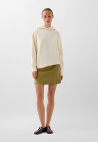 Cremefarvet hoodie med en frontlomme og hætte, parret med en tætsiddende olivengrøn nederdel. Modellen bærer sorte slip-on sko.