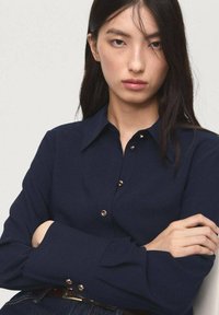Navy blue shirt with a button-up front, featuring gold-tone buttons and a classic collar. Long sleeves with buttoned cuffs. Smooth fabric texture.