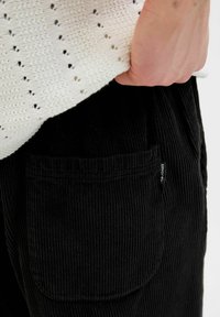 Sorte corduroy bukser med en frontlomme og vertikale riller. En creme strikbluse med åbne mønstre er delvist synlig.