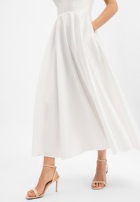 Robe longue blanche avec un corsage ajusté et une jupe fluide dotée de poches. Associée à des sandales à talons hauts nude à fines brides. Texture lisse.