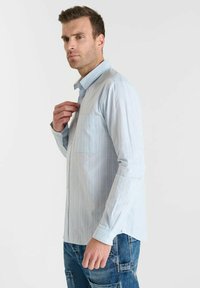 Man die de kraag van een lichtblauwe gestreepte overhemden met knopen aanpast, gekleed in blauwe denim cargobroek, staand tegen een effen lichte achtergrond.