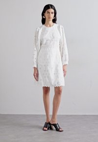 Robe blanche avec des motifs floraux brodés, décolleté festonné, longues manches bouffantes, et ourlet en dentelle festonnée. Talons ouverts noirs.