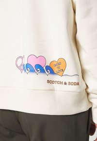 Cremefarbener Pullover mit Grafikherzen in Pink, Orange, blauen Wellen und dem Text "BORN TO LOVE", mit dem Markennamen "SCOTCH & SODA" am unteren Rand.