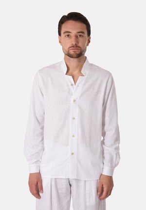 Uomo che indossa una camicia bianca a maniche lunghe con colletto alla mandarina e pantaloni bianchi abbinati, in piedi contro uno sfondo uniforme.