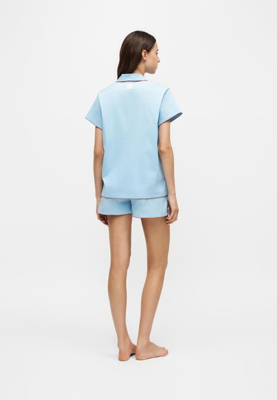 Femme debout pieds nus, de dos, portant un haut bleu clair à manches courtes et un short assorti avec un liseré rouge sur les bords.