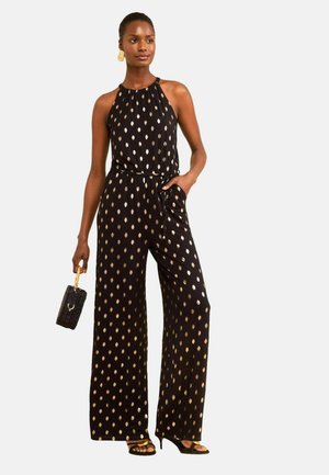 Kvinde iført sort halterneck-jumpsuit med gyldne polkaprikker, der holder en sort håndtaske og har sorte højhælede sko og gyldne øreringe på.