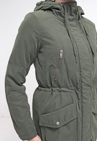Olivově zelená prošívaná bunda s kapucí, předním zipem, dvěma bočními kapsami a stahováním v pase. Texturovaná látka s jemným leskem.