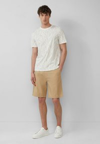 Witte herenoverhemd met een subtiel bladerenpatroon, gecombineerd met beige shorts. Schoeisel is witte sneakers. Schoon ontwerp, casual fit.