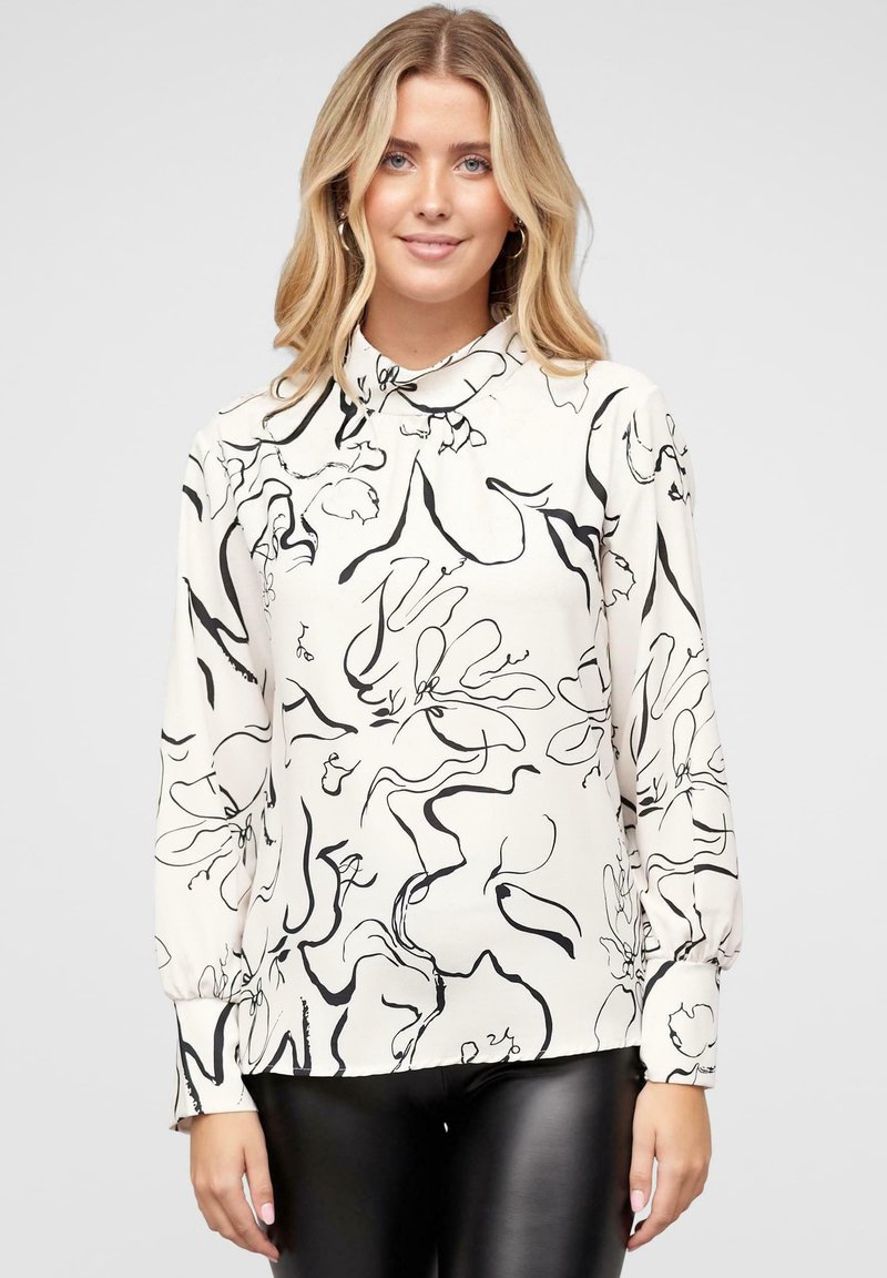 Blouse crème avec un motif floral abstrait noir, à manches longues et col haut. Tissu doux avec une texture lisse et un léger éclat.