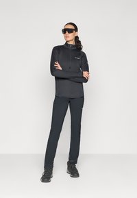 Egy nő áll keresztbe tett karokkal, fekete szabadtéri dzsekit, nadrágot, túracipőt és oversize fekete napszemüveget visel egy egyszínű háttér előtt.