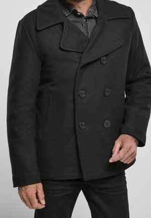 Mann trägt einen schwarzen doppeltgeknöpften Pea Coat über einem dunkelblauen Jeanshemd, Hände sichtbar, neutraler grauer Hintergrund.