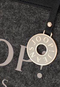 Rund guld- och grå nyckelring med texten "JOOP! JEANS" fäst vid en strukturerad grå tygyta med stora tryckta bokstäver.