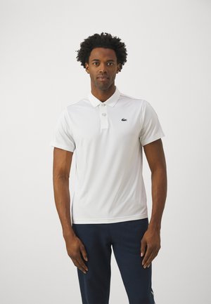 Polo shirt blanc à manches courtes, avec patte de boutonnage à trois boutons et logo brodé vert ; associé à un pantalon bleu foncé. Texture de tissu lisse.