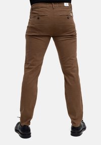 Chinos in cotone marrone con vestibilità slim, dotati di tasche posteriori con chiusura a bottone e un'etichetta sulla vita. Calzature nere visibili sotto.