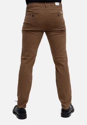 Chinos in cotone marrone con vestibilità slim, dotati di tasche posteriori con chiusura a bottone e un'etichetta sulla vita. Calzature nere visibili sotto.