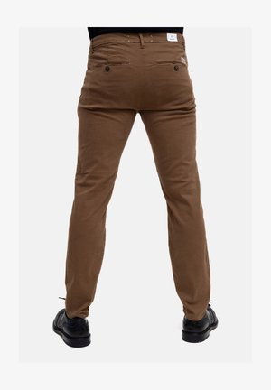 Chinos in cotone marrone con vestibilità slim, dotati di tasche posteriori con chiusura a bottone e un'etichetta sulla vita. Calzature nere visibili sotto.