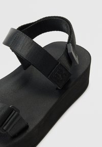Juoda platforminė sandalai su plačiomis tekstilėmis dirželių ir tekstūruota guminėmis padais. Yra stačiakampis vidpadis ir logotipo detalė ant dirželio.