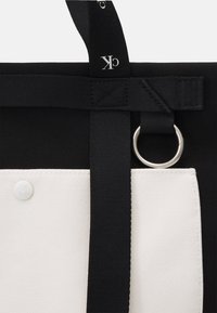 Borsa tote bianco e nero con tessuto strutturato, caratterizzata da una grande tasca bianca, tracolle nere e un anello metallico argento come dettaglio.